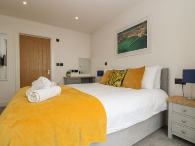 A bedroom with a double bed with yellow cushions and blanket white towels on bed grey bedside tables with lamps and a wooden door at Valley View in Mullion