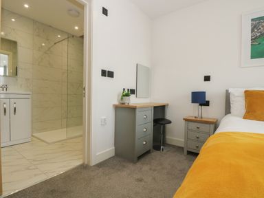 A bedroom with a bed having a yellow blanket a bedside table with a blue lamp a dressing table with a mirror and stool and an ensuite bathroom with a shower at Valley View in Mullion