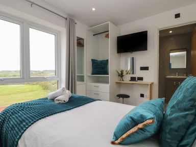A bedroom with a bed, teal pillows and blanket, a window with curtains, a wardrobe, a wall-mounted TV, and a doorway to a bathroom at Valley View in Mullion