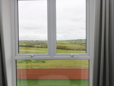 A window with curtains showing a view of green fields and a wooden fence at Valley View in Mullion