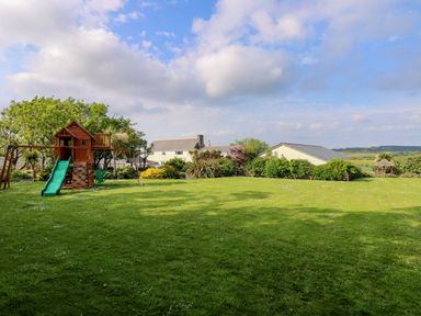 A large grassy garden area with a wooden play structure and houses in the background at Valley View in Mullion