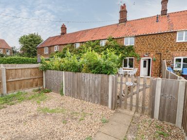 A fenced garden with gravel and a wooden gate in front of a brick house with red roof tiles at Tawny Cottage in Heacham