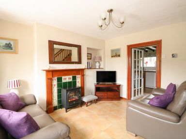 A living room with two sofas facing each other a fireplace with green tiles and a small stove a wooden TV stand with a TV and a small upholstered footstool at Tawny Cottage in Heacham