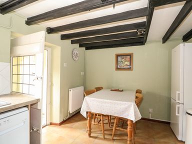 A dining area with a table covered by a cloth and four wooden chairs a wall clock a framed picture and a refrigerator at Tawny Cottage in Heacham