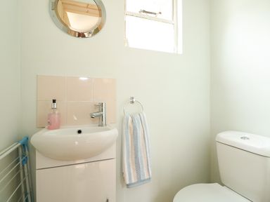 A small bathroom with a white sink, a pink soap dispenser, a round mirror, a towel ring with a striped towel, and a white toilet at Tawny Cottage in Heacham