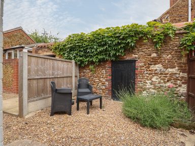 A small outdoor patio area with two black chairs a table gravel ground and a stone wall with greenery at Tawny Cottage in Heacham