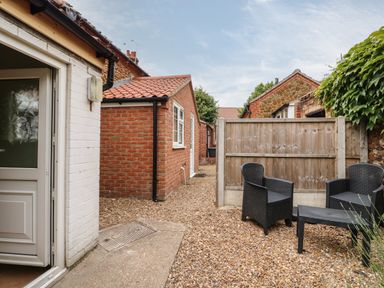 A small outdoor area with gravel ground two black chairs and a table next to brick and white painted walls at Tawny Cottage in Heacham