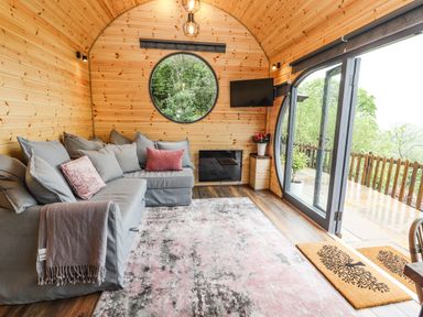 A wooden room with a grey sectional sofa and cushions a circular window a wall-mounted tv and a glass door leading outside at Offas Dyke Escape in St Asaph