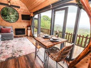 A dining table with chairs and tableware next to large windows with a countryside view in a wooden room with a sofa and a round window at Offas Dyke Escape in St Asaph