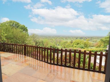 A patio with wet stone tiles and a wooden railing overlooking a green landscape with trees and fields at Offas Dyke Escape in St Asaph