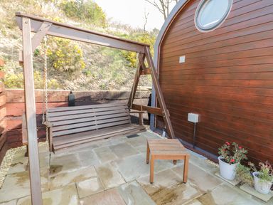 An outdoor patio area with a wooden swing bench a small wooden table and potted plants at Offas Dyke Escape in St Asaph