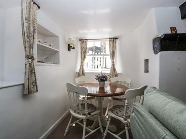 A small dining area with a round wooden table and four white chairs near a window with floral curtains at Small Piece in Porlock