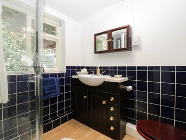 A bathroom with a sink cabinet dark blue wall tiles a wooden toilet seat and a window at Small Piece in Porlock