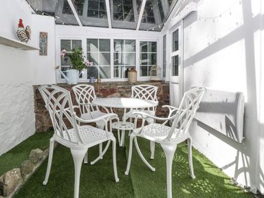 A sunroom with white metal table and chairs on artificial grass with a watering can and plants on a stone ledge at Small Piece Porlock
