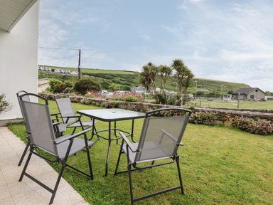 An outdoor patio with a table and four chairs on grass with bushes hills and houses in the background at 7 Europa Court in Mawgan Porth