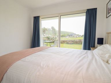 A bedroom with a bed covered in white linens next to sliding glass doors with blue curtains showing a garden and a house outside at 7 Europa Court in Mawgan Porth