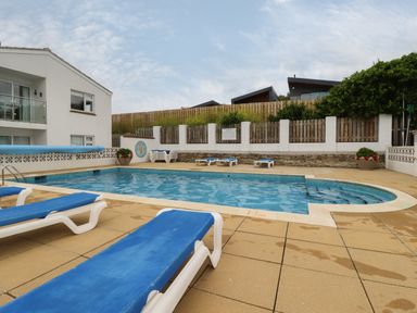 An outdoor swimming pool with blue lounge chairs and a white building nearby at 7 Europa Court in Mawgan Porth
