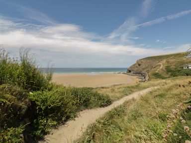 A sandy beach with a walking path through grass leading to the shore and cliffs in the background at 7 Europa Court in Mawgan Porth