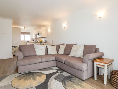 A living room with a brown corner sofa and a rug with circular patterns adjacent to a kitchen at 7 Europa Court in Mawgan Porth
