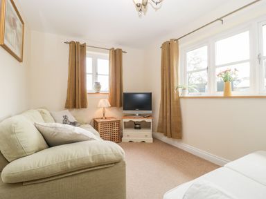 A small living room with a beige sofa cushions a lamp television and multiple windows with brown curtains at The Coal House at Mendip View in Wedmore