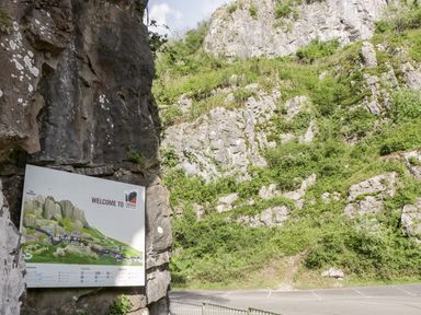 A sign welcoming visitors to Cheddar Gorge and Caves near rocky cliffs and a parking area