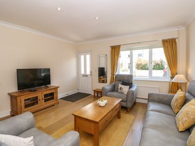 A living room with a television on a wooden stand a coffee table two armchairs a sofa and a window with yellow curtains at Inglewood in Keswick
