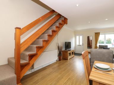 A living room with wooden stairs carpeted in beige a wooden dining table with plates and cutlery a TV on a wooden cabinet and a window with yellow curtains at Inglewood in Keswick