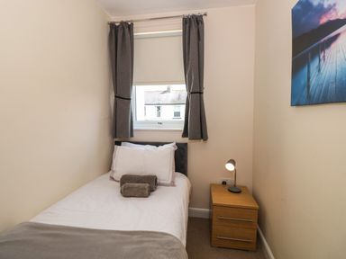 A single bed with white bedding and brown towels next to a window with grey curtains and a wooden bedside table with a lamp in a small room at Inglewood in Keswick