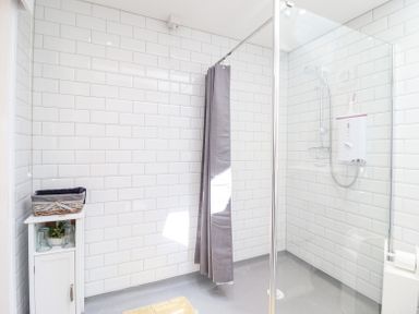 A bathroom with white tiled walls a shower area with a gray curtain a white cabinet holding a basket and glasses and a showerhead with a water heater at Varley Villa in Newtown