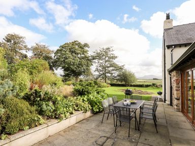 A patio with a dining table and chairs next to a garden with trees and bushes at Clynnog House in Dwyran near Newborough