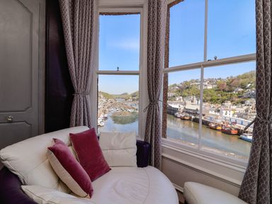 A seating area with white and purple cushions next to large windows overlooking a harbor with boats and hillside houses at Albany in Looe