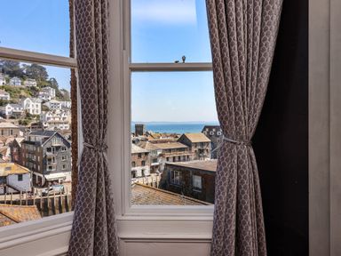 A window with patterned curtains showing rooftops and a distant sea view at Albany in Looe