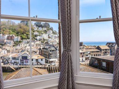 A view through a window with curtains showing houses on a hillside and the sea beyond at Albany in Looe