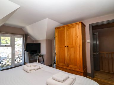 A bedroom with a bed, folded towels, wooden wardrobe, desk with a chair and a television near a window at Albany in Looe