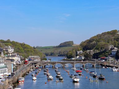 A river with moored boats and a stone bridge connecting banks with buildings and hills in the background at Albany in Looe