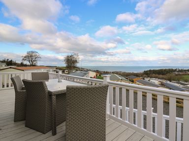 An outdoor deck with a table and four chairs overlooking houses trees and the sea at McAuliffe Lodge in Borth