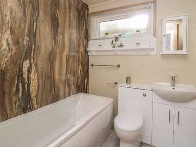 A bathroom with a bathtub and marble wall panel a toilet and sink with cabinet under a frosted window with small plants and a fish ornament on the ledge at McAuliffe Lodge in Borth
