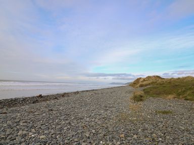 A pebble beach with grassy sand dunes and sea waves under a partly cloudy sky at McAuliffe Lodge in Borth