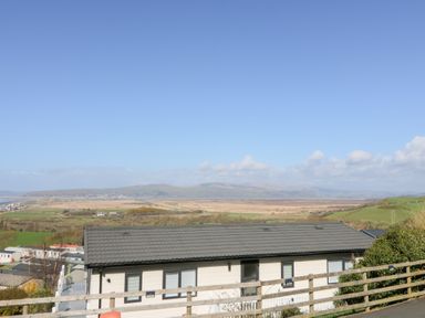 A house with a grey roof and multiple windows with a wooden fence in front and a landscape of fields and hills under a blue sky at McAuliffe Lodge in Borth
