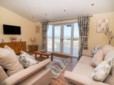 A living room with two sofas a coffee table and TV facing balcony doors with sea view at McAuliffe Lodge in Borth