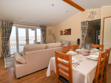 A dining table set for six next to a beige sofa in a living room with glass doors and patterned curtains at McAuliffe Lodge in Borth