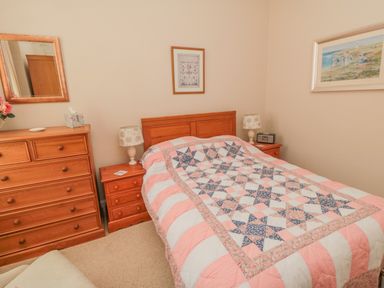 A bedroom with a double bed with a patterned quilt two wooden bedside tables lamps a wooden chest of drawers and framed pictures on cream walls at Sandsend Sunset View in Whitby