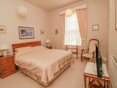 A bedroom with a bed nightstands lamps paintings a window with curtains an armchair and a television on a table at Sandsend Sunset View in Whitby