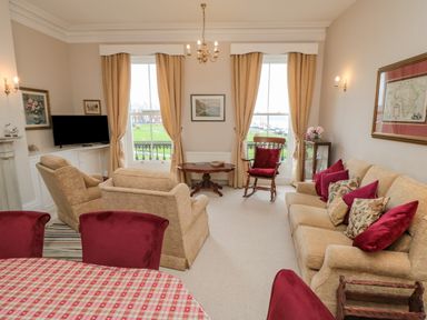 A living room with beige sofas and armchairs red cushions and curtains and a dining table with red chairs at Sandsend Sunset View Whitby