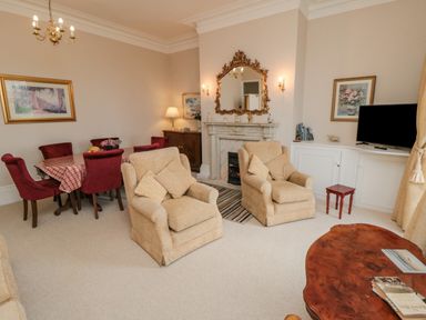 A living room with two beige armchairs a wooden coffee table a dining table with red chairs and a television at Sandsend Sunset View in Whitby