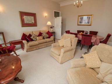A living room with beige sofas and armchairs a wooden dining table with red chairs and patterned tablecloth at Sandsend Sunset View Whitby
