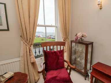 A wooden chair with red cushions in front of a window with beige curtains next to a glass cabinet with glassware and a small wooden table with flowers at Sandsend Sunset View Whitby
