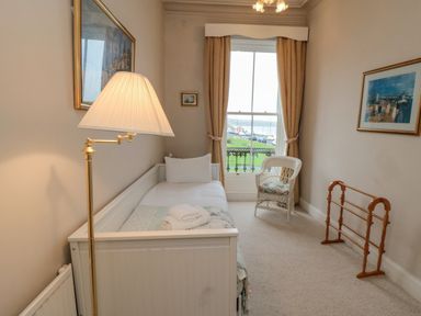A small bedroom with a single white daybed a floor lamp a wicker chair and framed pictures at Sandsend Sunset View in Whitby