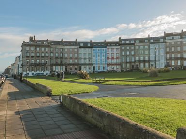 A row of multi-story terraced houses facing a green park with benches and a paved walkway at Sandsend Sunset View in Whitby