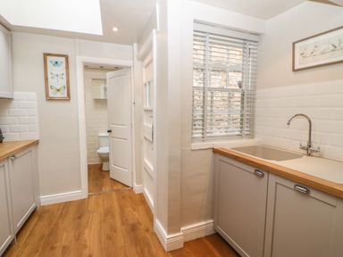 A kitchen with wooden floor cabinets a sink and a window with blinds showing a small bathroom in the background at Chapel Cottage in Corbridge